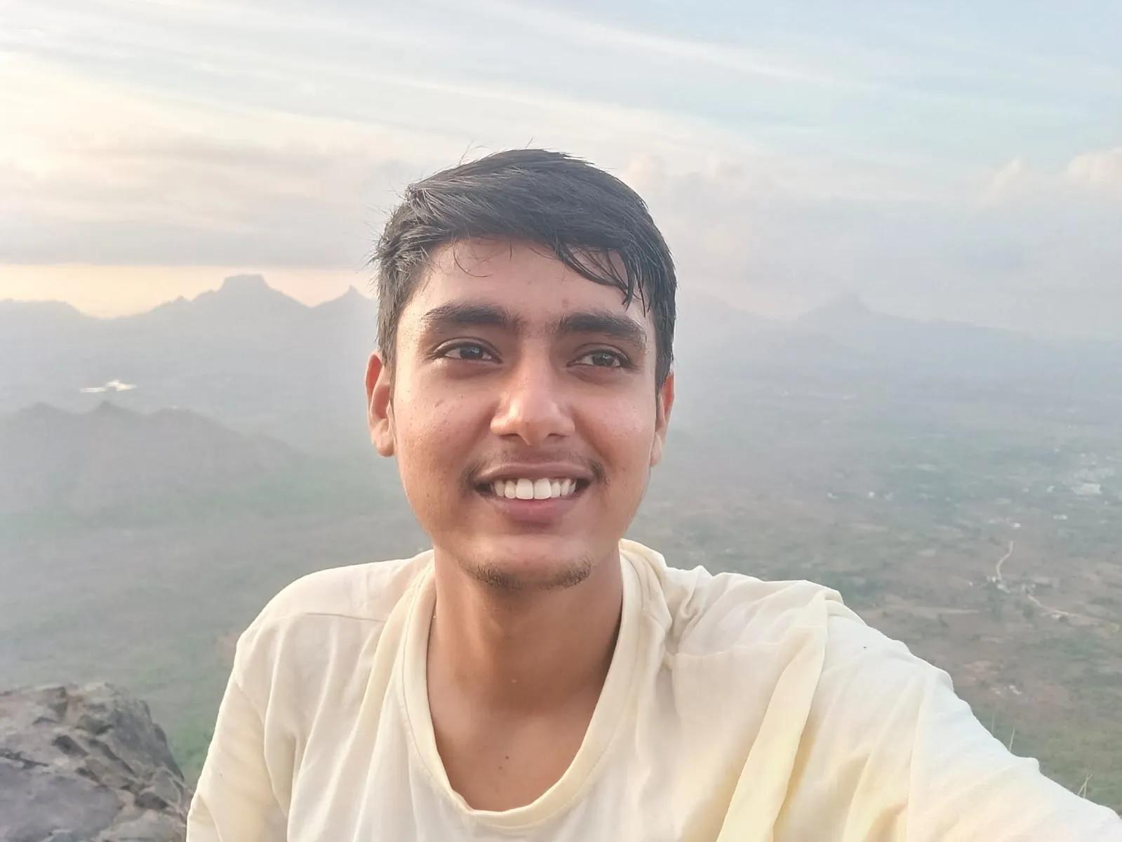 Kartik smiling on a hilltop with mountains behind him